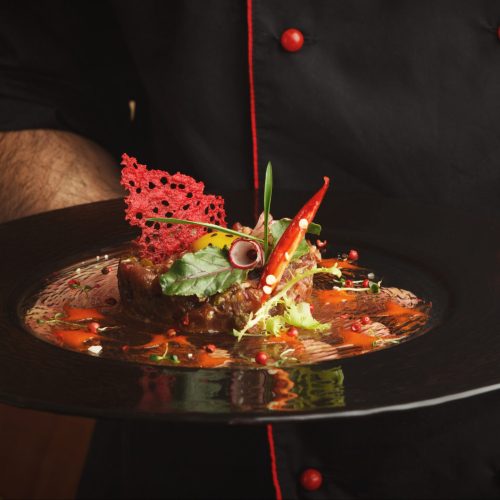 Male chef offering exclusive restaurant food. Traditional appetizer - raw veal tartare with sauce, quail egg, greens, vegetables and crisp on black plate. Organic healthy meals in modern serving