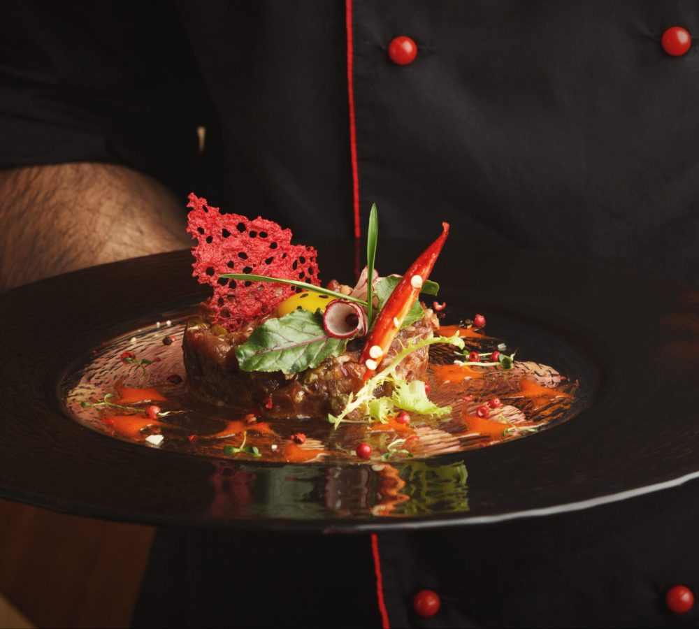 Male chef offering exclusive restaurant food. Traditional appetizer - raw veal tartare with sauce, quail egg, greens, vegetables and crisp on black plate. Organic healthy meals in modern serving